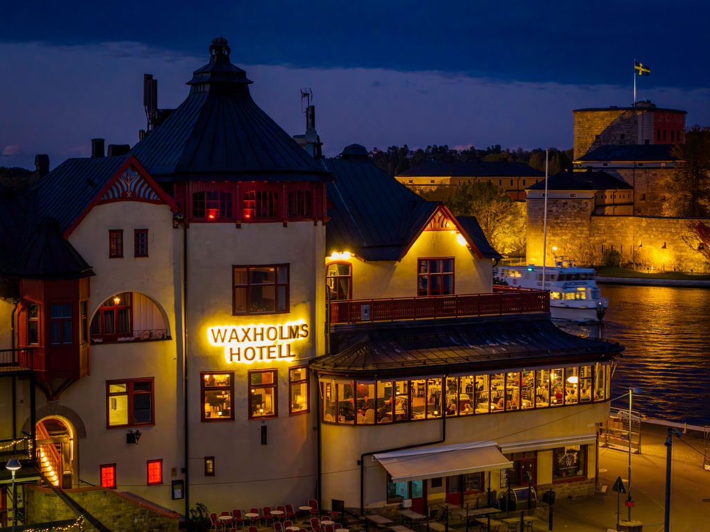 Waxholms Hotell upplyst i kvällsblått med Vaxholms kastell i bakgrunden
